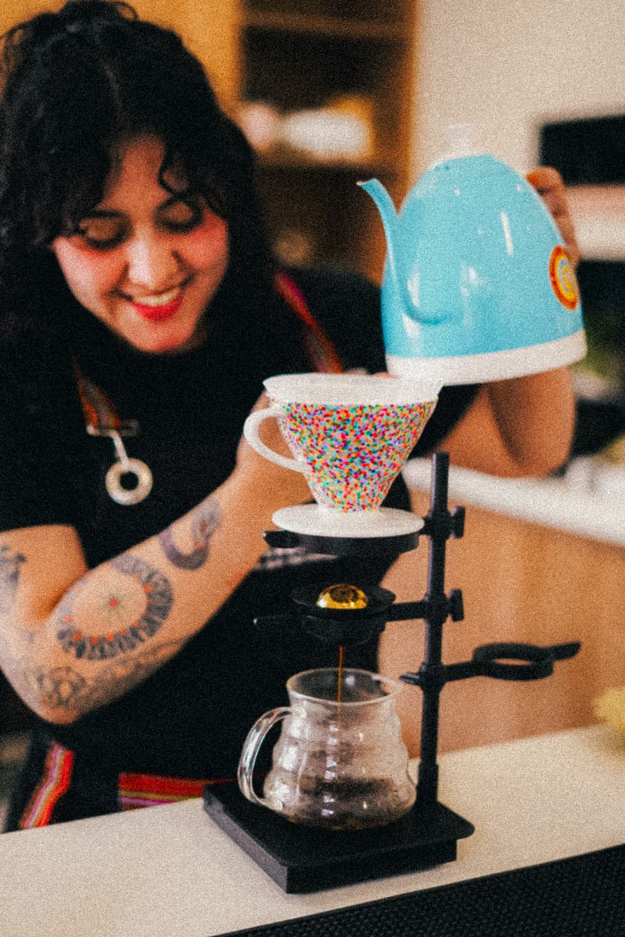 Mujer sonriente vertiendo agua de una tetera azul en un filtro de café con motas coloridas.