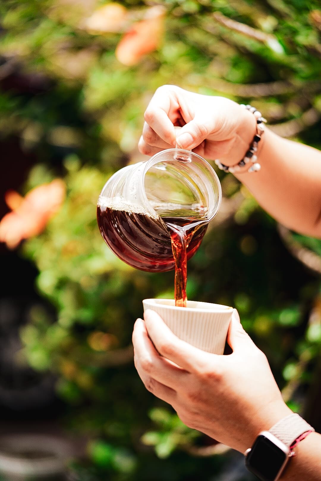 Persona vertiendo café oscuro de una jarra de cristal a una taza blanca entre vegetación.