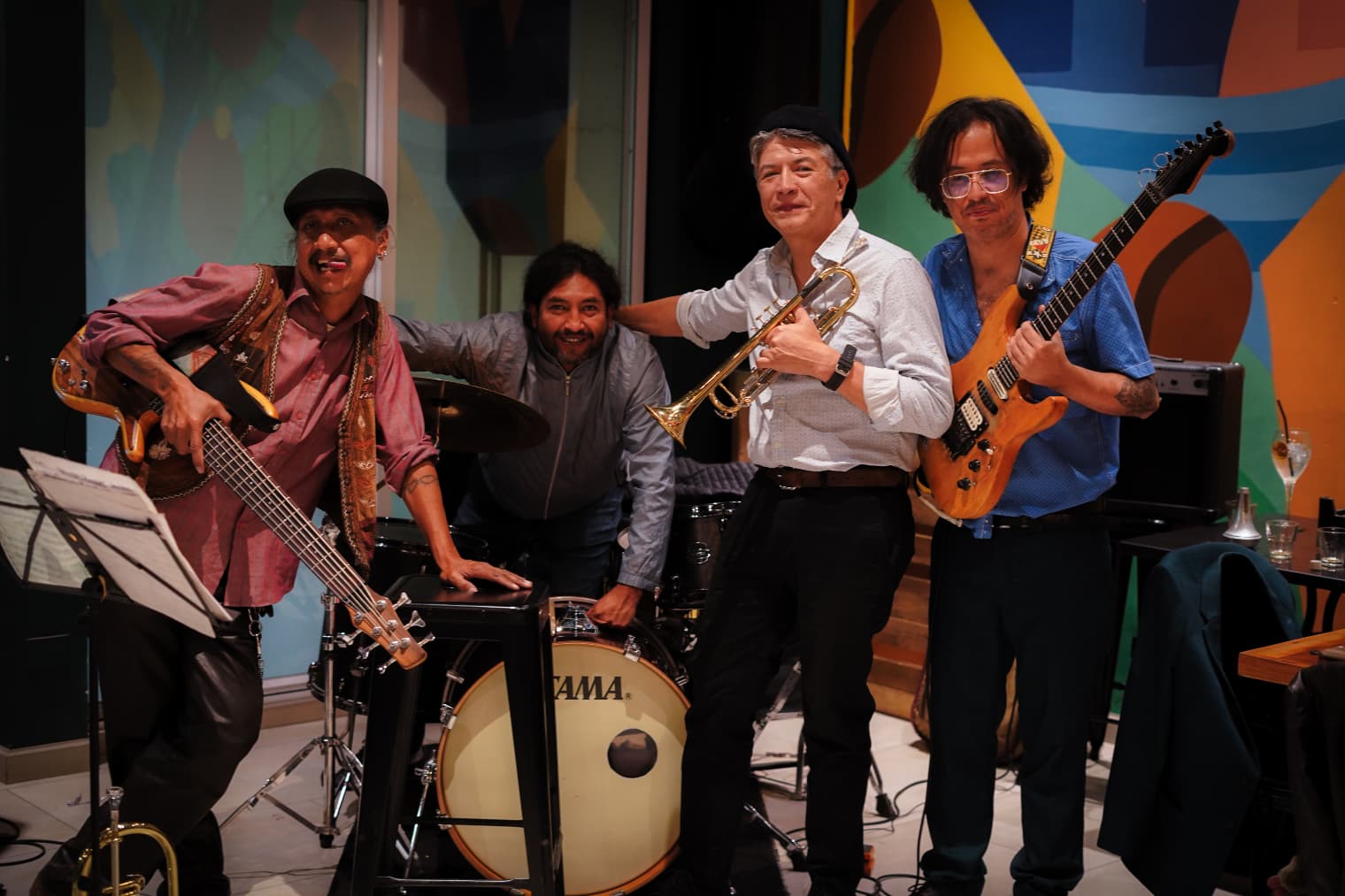 Cuatro músicos sonrientes posan con bajo, batería, trompeta y guitarra eléctrica frente a un mural.