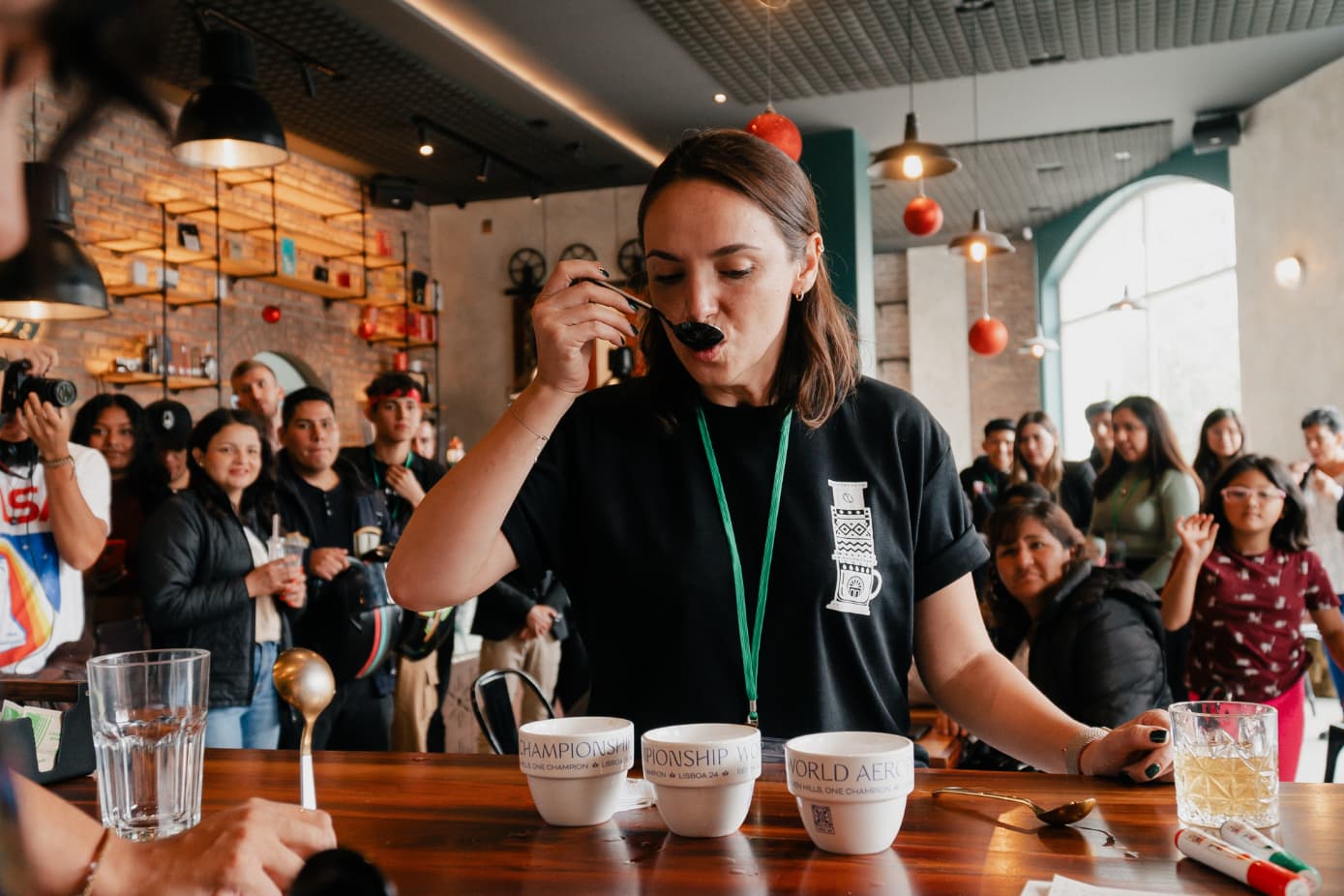 Mujer catando café con cuchara en una competencia, rodeada de espectadores en una cafetería moderna.