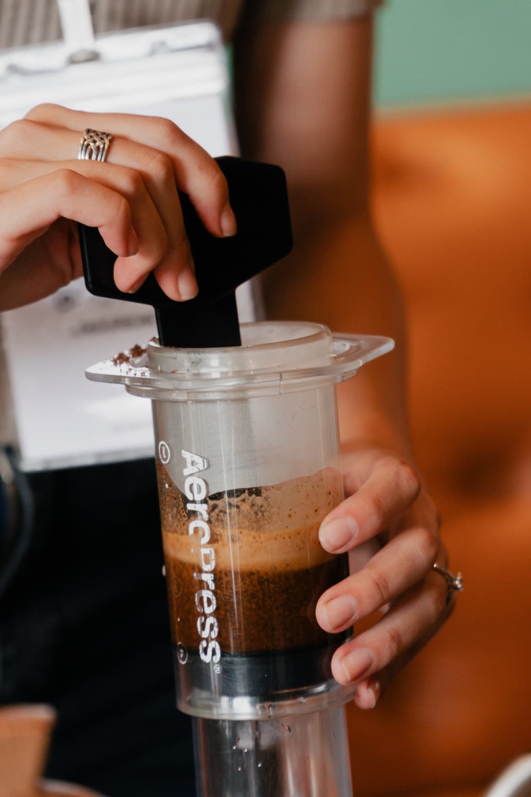 Manos revolviendo café molido con una paleta negra dentro de una cafetera AeroPress transparente.